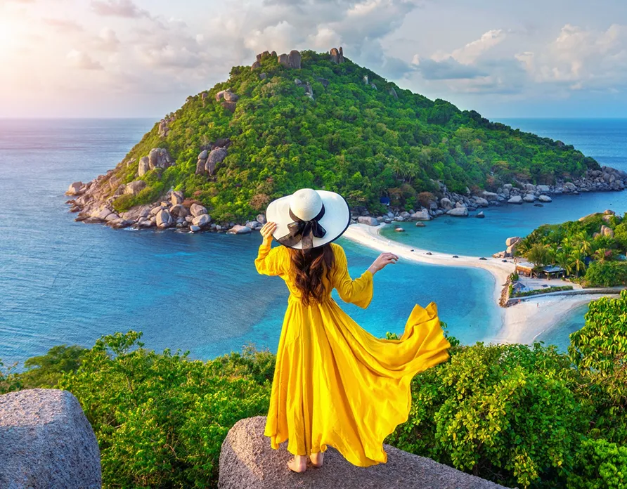 Woman in a flowing yellow dress and large hat stands on a rock overlooking a green island and blue sea.