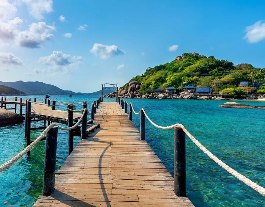 Wooden pier with ropes leading to a green island over clear turquoise sea under a blue sky.